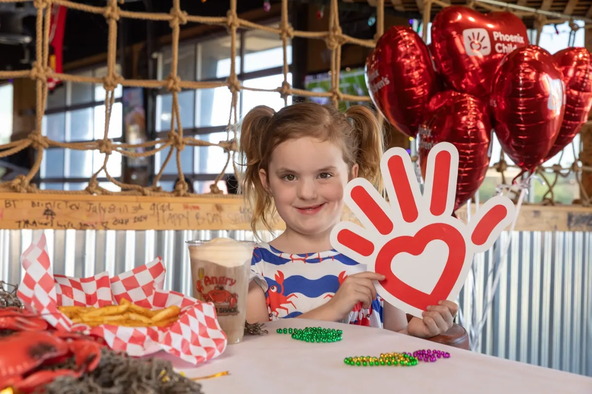 A smiling girl at an Angry Crab Shack table holding up the Phoenix Children’s hand sign